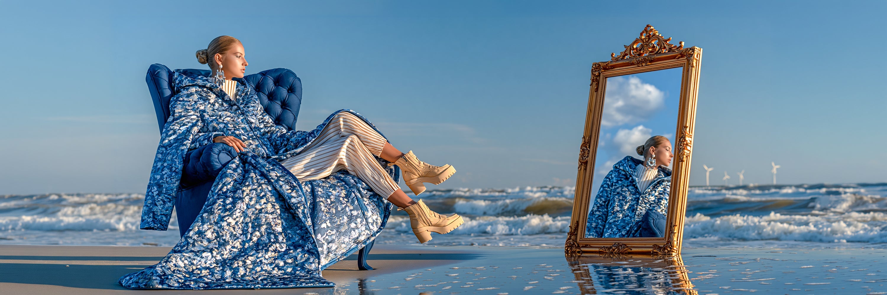 Selbstbewusste Frau auf einem grossen blauen Sessel am Strand neben einem prunkvollen goldfarbenen Spiegel, der sie reflektiert, während sie auffällige silberfarbene Ohrringe trägt.