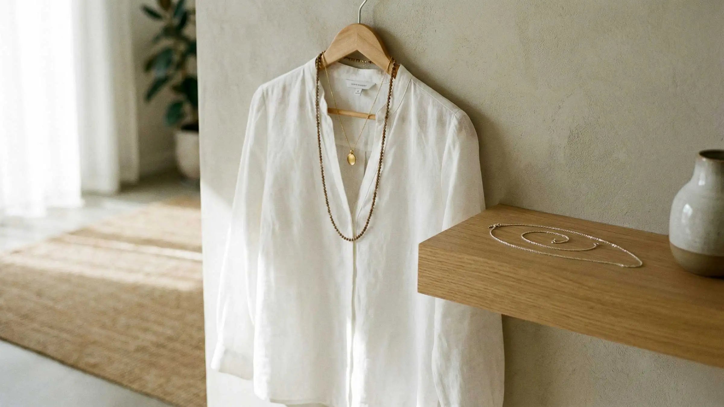 Weisse Bluse am Bügel mit Layering-Ketten im warmen Tageslicht, daneben Schmuck auf Holzablage, Avirelle.