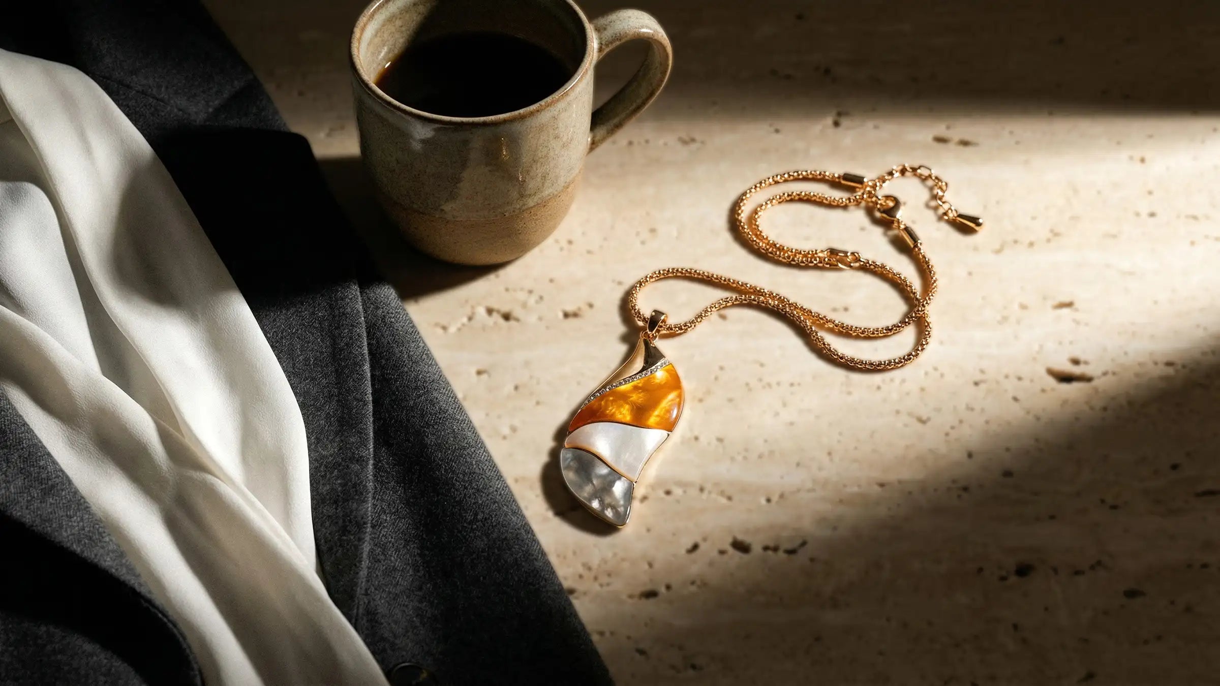 Collier mit Perlmutt-Anhänger auf Travertin neben Bluse, Blazer und Kaffeetasse im Fensterlicht, Avirelle.